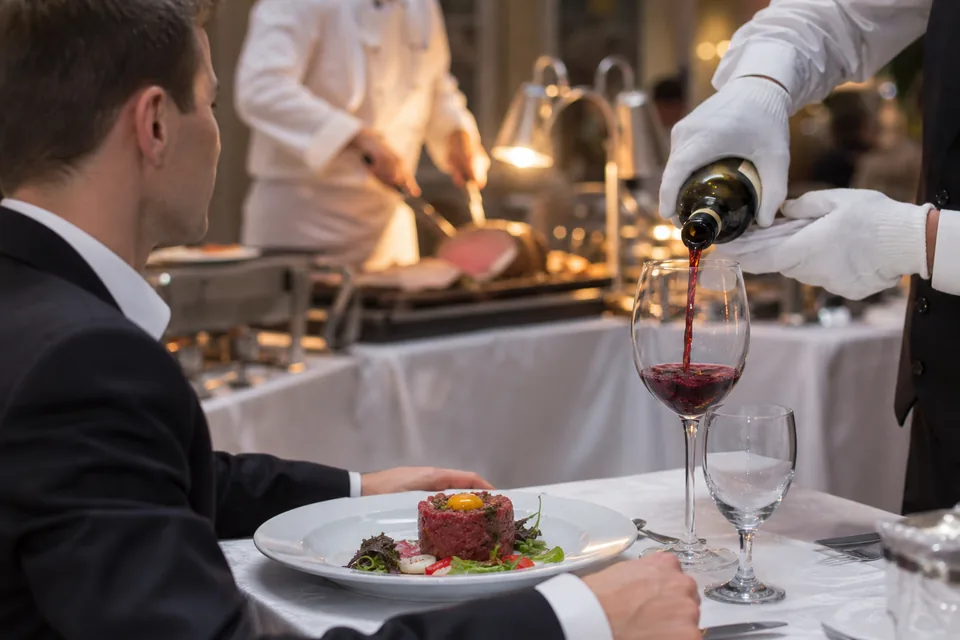 Menu bankietowe na stole, przystawka na białym talerzu, kelner nalewa wino, w tle bufet z daniami w podgrzewaczach