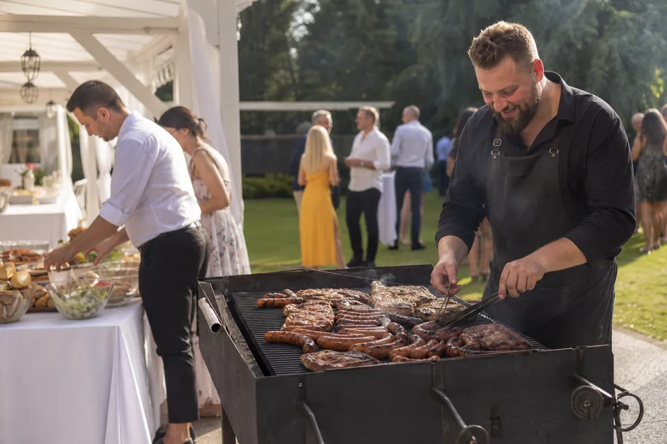 Grill na poprawinach, grillmistrz przy ruszcie i kelner uzupełniający bufet z sałatkami pod pawilonem w ogrodzie